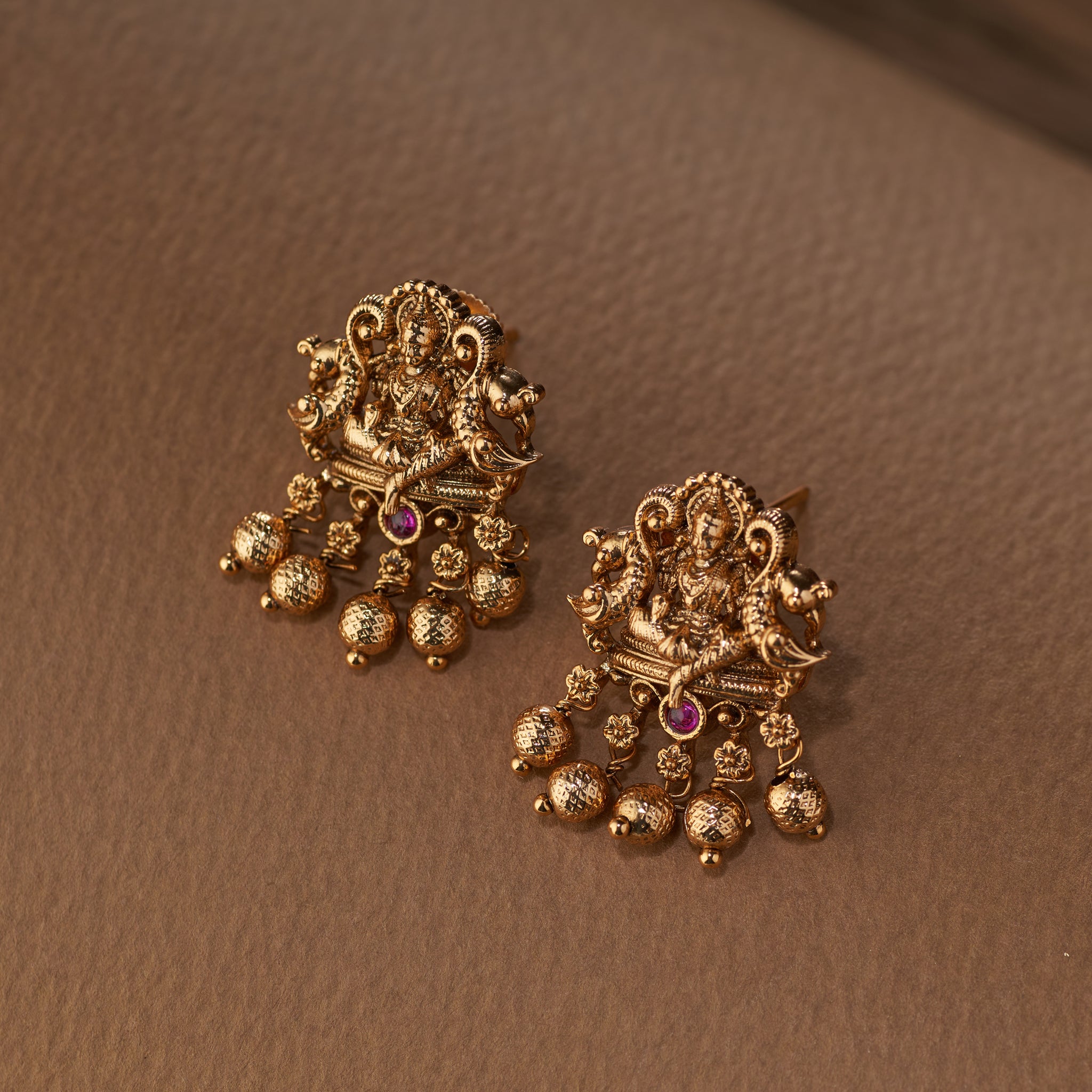 A pair of Tilloma antique gold temple earrings, with a detailed depiction of the goddess Lakshmi and peacocks and hanging gold beads, photographed on a light brown surface.