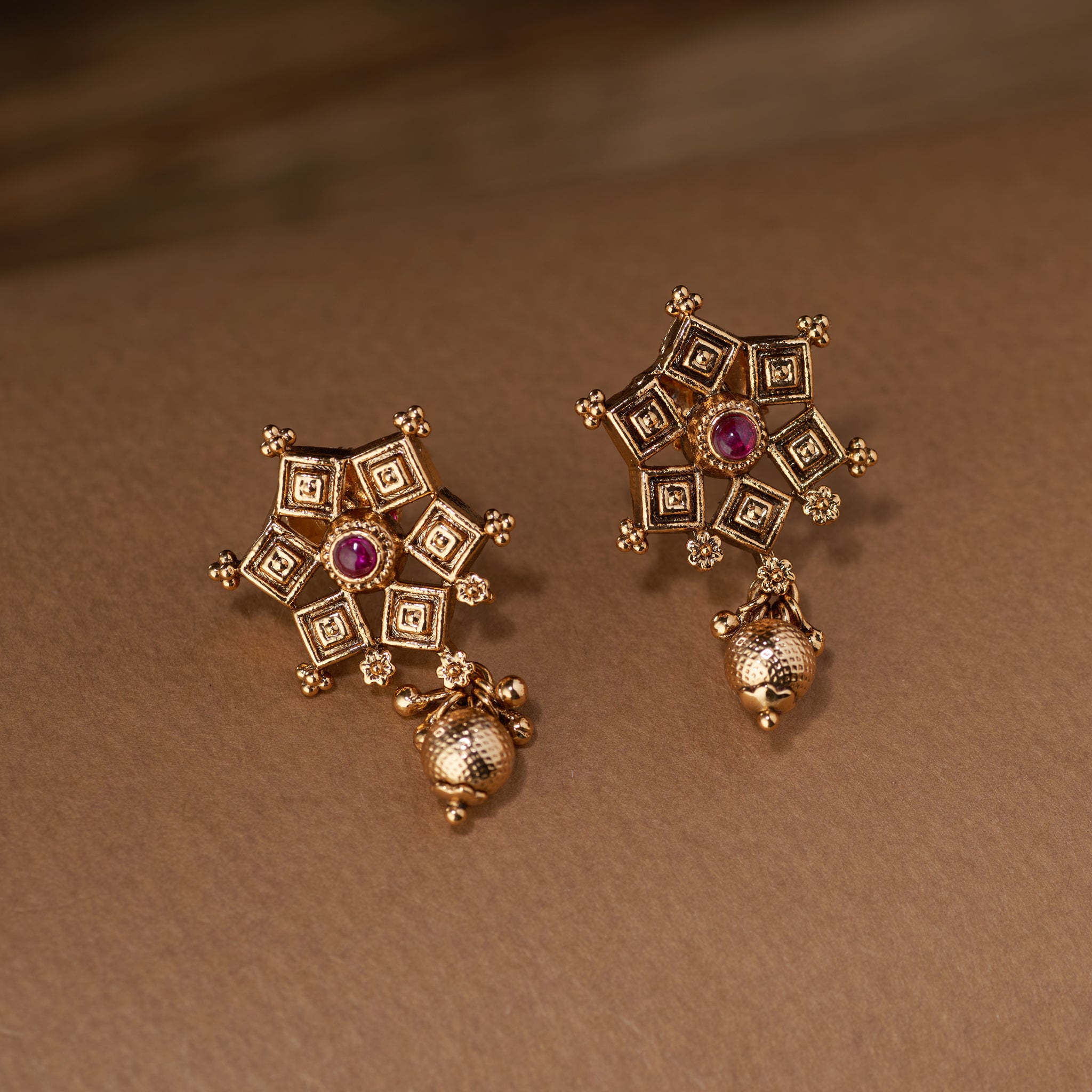 A pair of antique gold temple jewellery earrings with a star-shaped design, set with a central ruby stone and a textured gold bead drop.