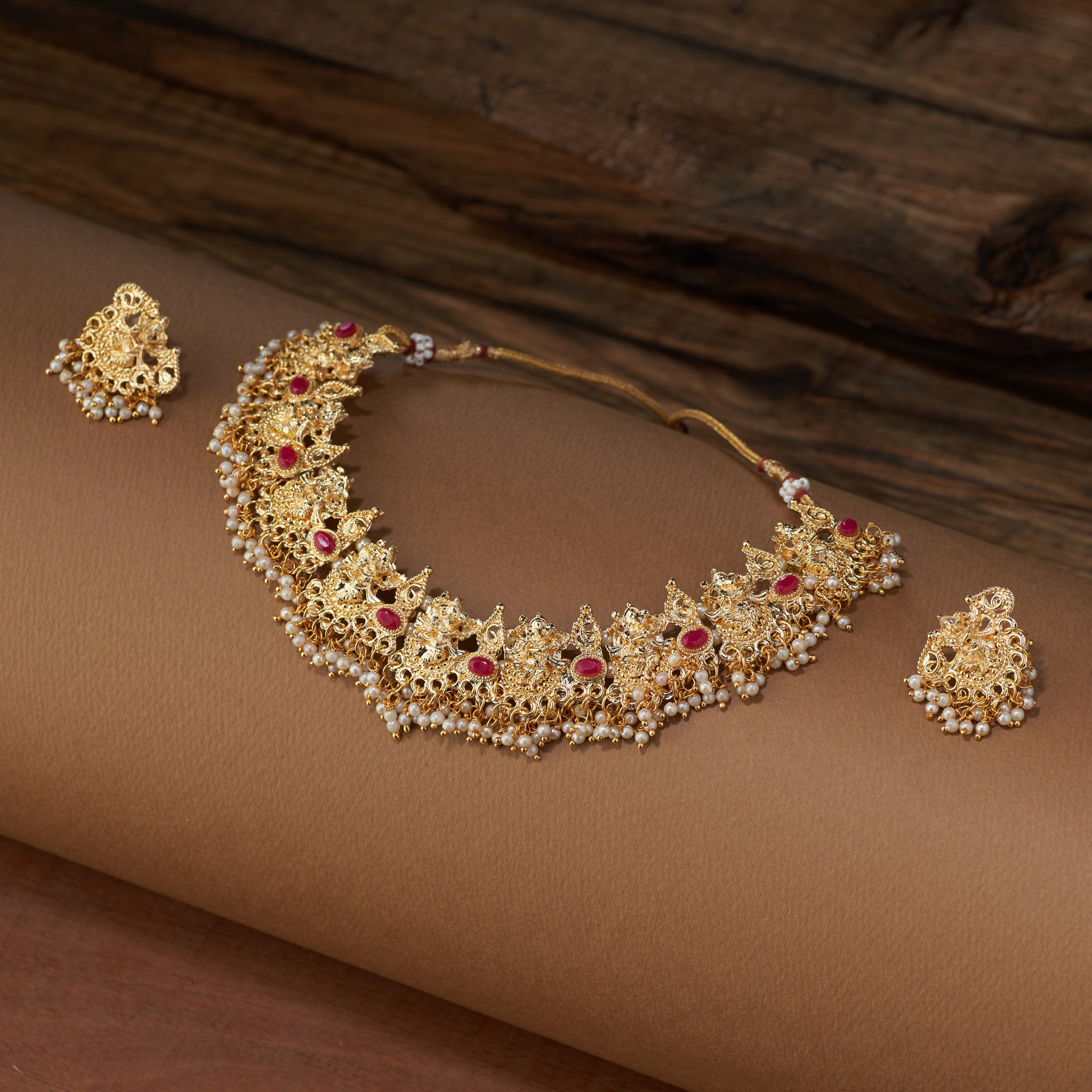 A full Ganesha temple jewelry set, including a gold-plated necklace and matching earrings with ruby stones and pearl beads, laid out on a brown background.
