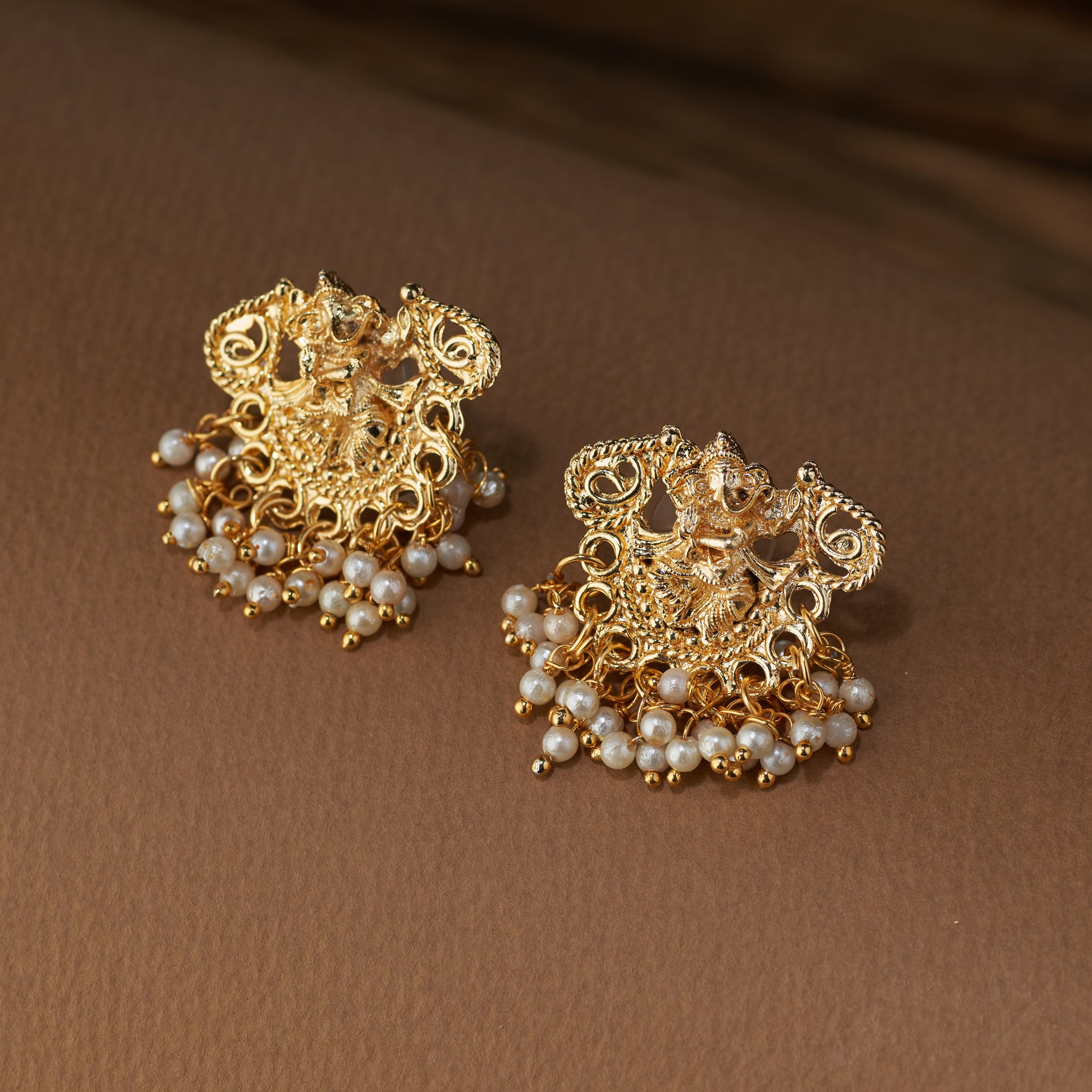 A pair of gold-plated Ganesha temple earrings, featuring intricate detail, a Ganesha motif, and pearl bead dangles on a brown surface.