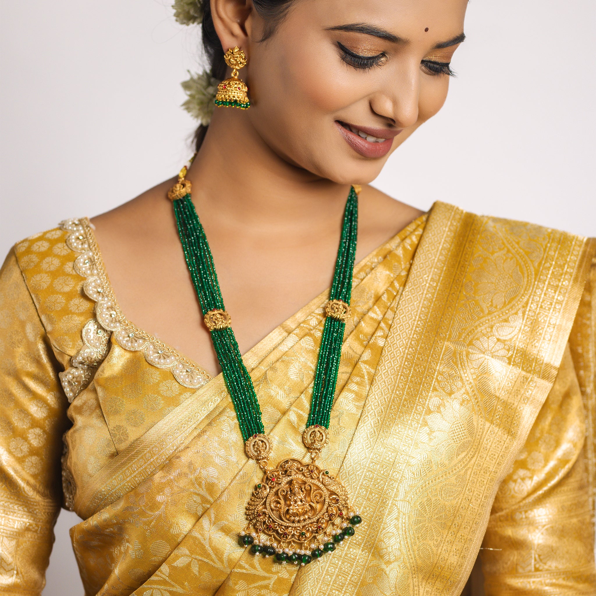 Woman wearing a gold saree with an emerald green South Indian Temple necklace and earrings on a plain background