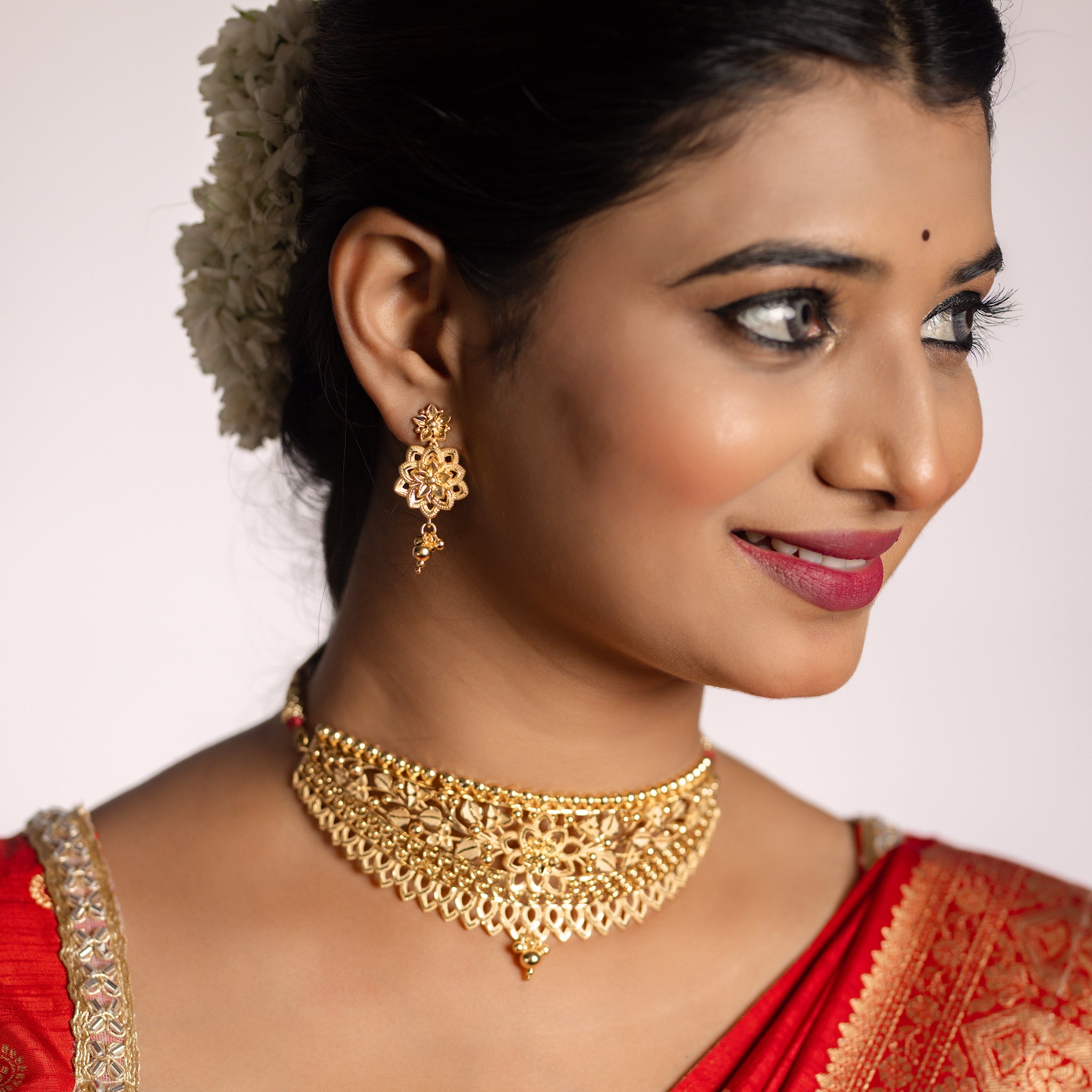 Woman wearing gold jewellery including earrings and a choker necklace, with a red garment. Gold Choker, forming jewellery, Poovi by Tilloma