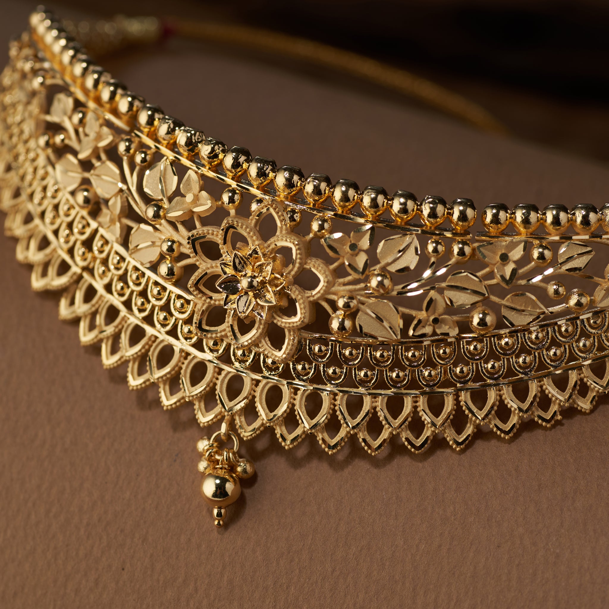 Gold necklace with intricate design on a brown background. Gold choker jewellery set with necklace, earrings on a brown surface. Gold Choker, forming jewellery, Poovi by Tilloma.