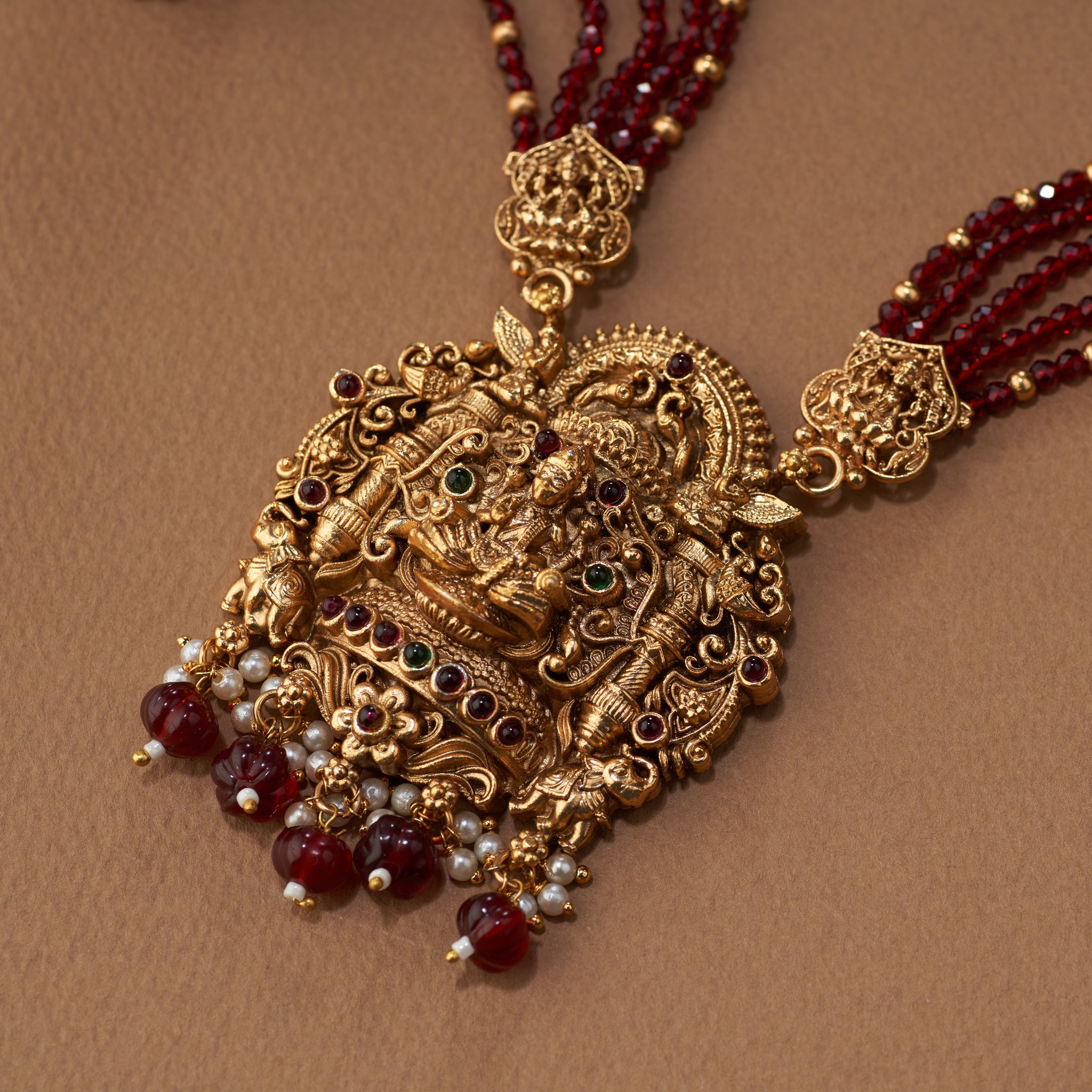 Ornate gold necklace with marron beads on a brown background. Gold and maroon beaded necklace with matching earrings on a brown background. Maroon beaded necklace with gold Lakshmi pendants, South Indian Temple jewellery on a brown background. matt gold finish and paired with earrings.