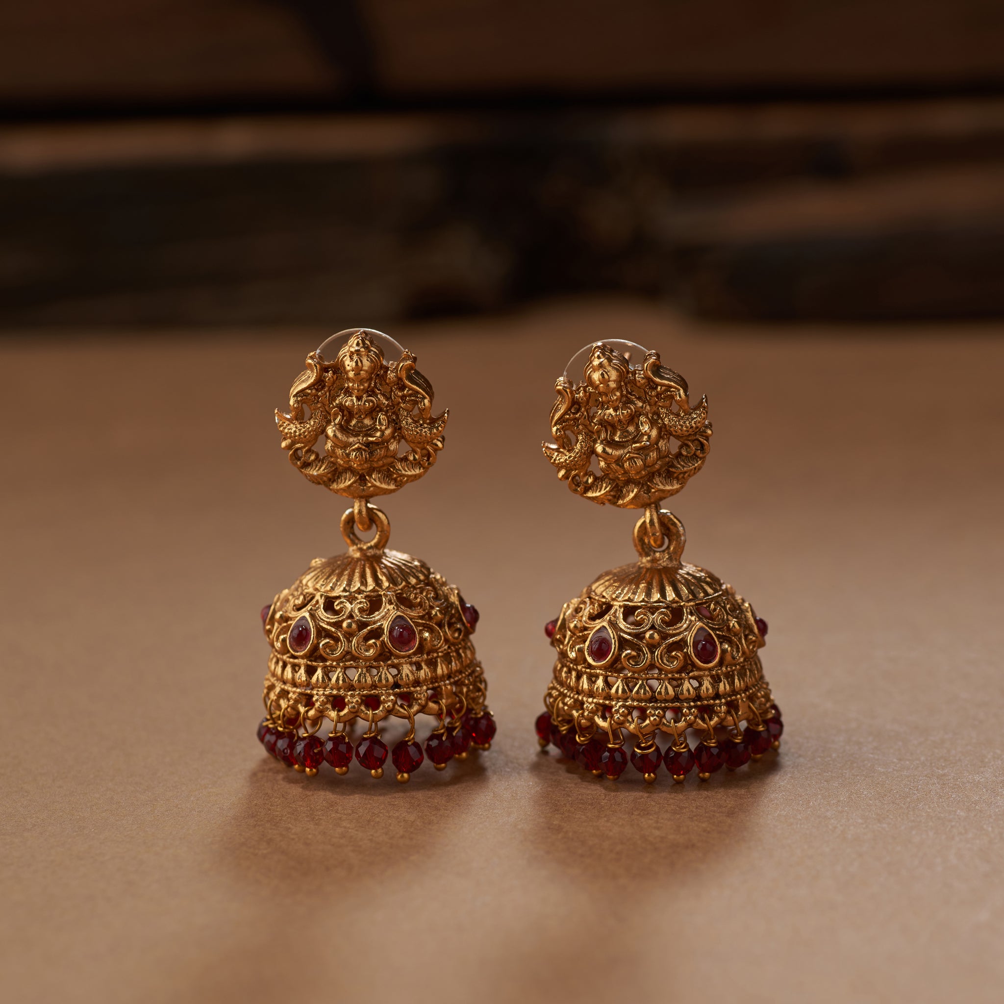 Pair of gold earrings with red gemstones on a brown background. Gold necklace with red beads and tassels on a brown background. Maroon beaded gold Lakshmi pendant intricate design South Indian Temple jewellery necklace set with matching jumkhis or earrings. Matt gold plated.