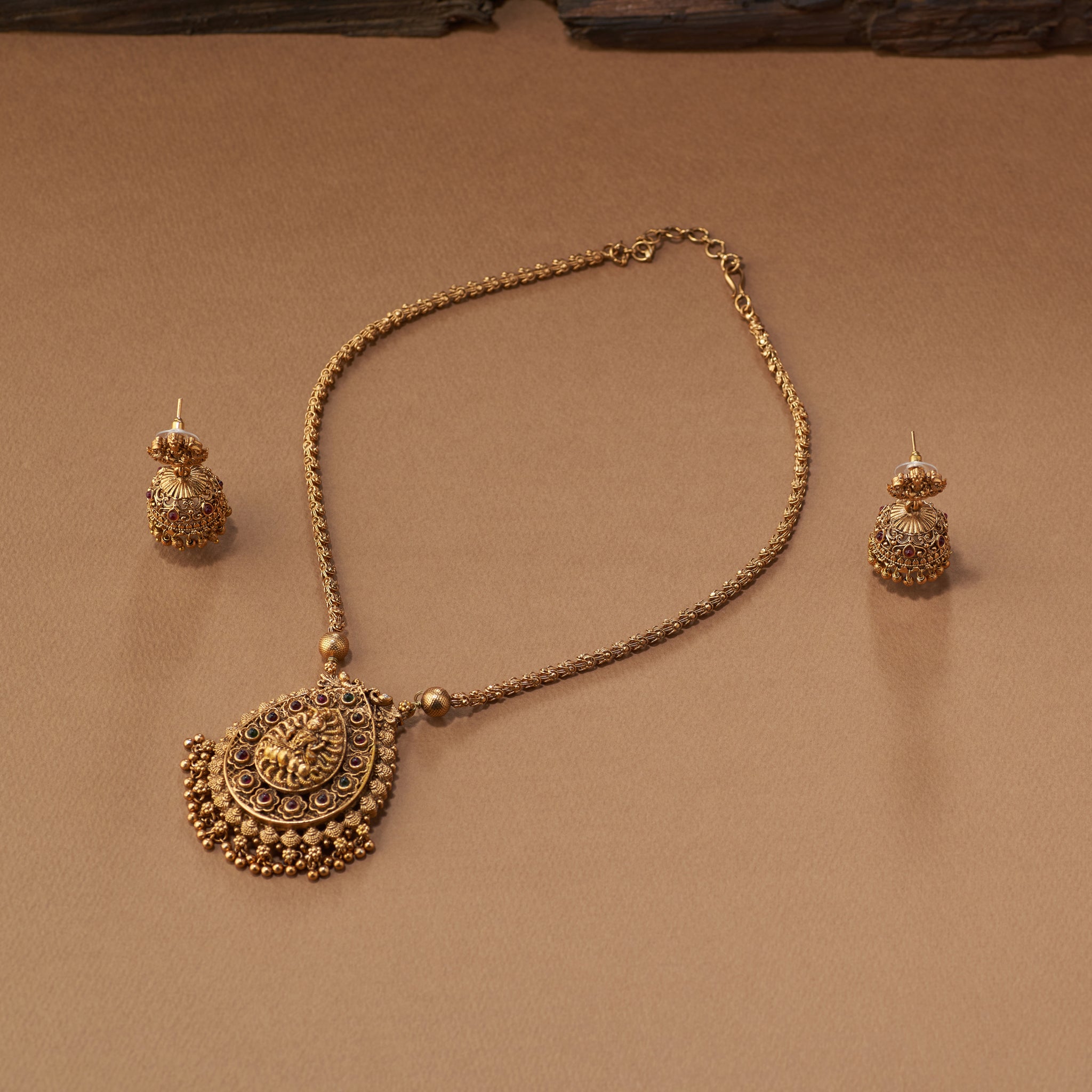 Gold necklace and earrings on a brown background. Woman wearing a gold saree with matching south Indian Temple jewelry with Lakshmi Motif. Temple Jewellery Lakshmi | Tilloma. Matt gold.
