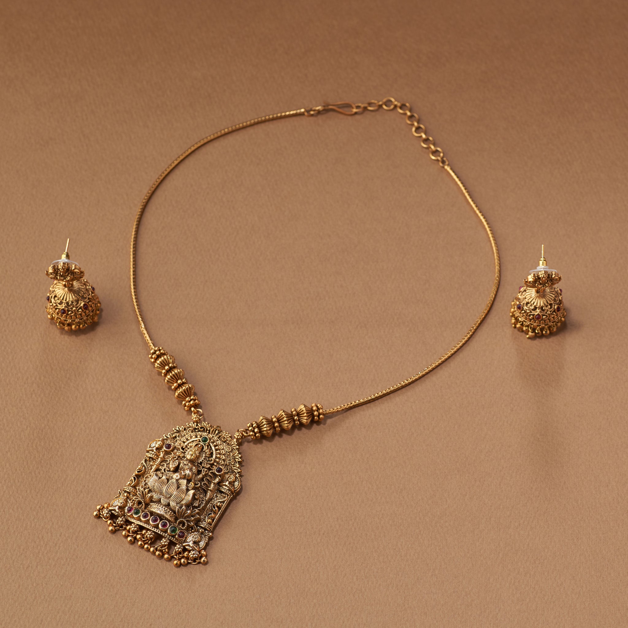 Gold necklace and earrings on a brown background. South Indian Temple jewelry with Lakshmi Motif. Temple Jewellery Lakshmi | Tilloma. Matt gold with intricate design on a brown background