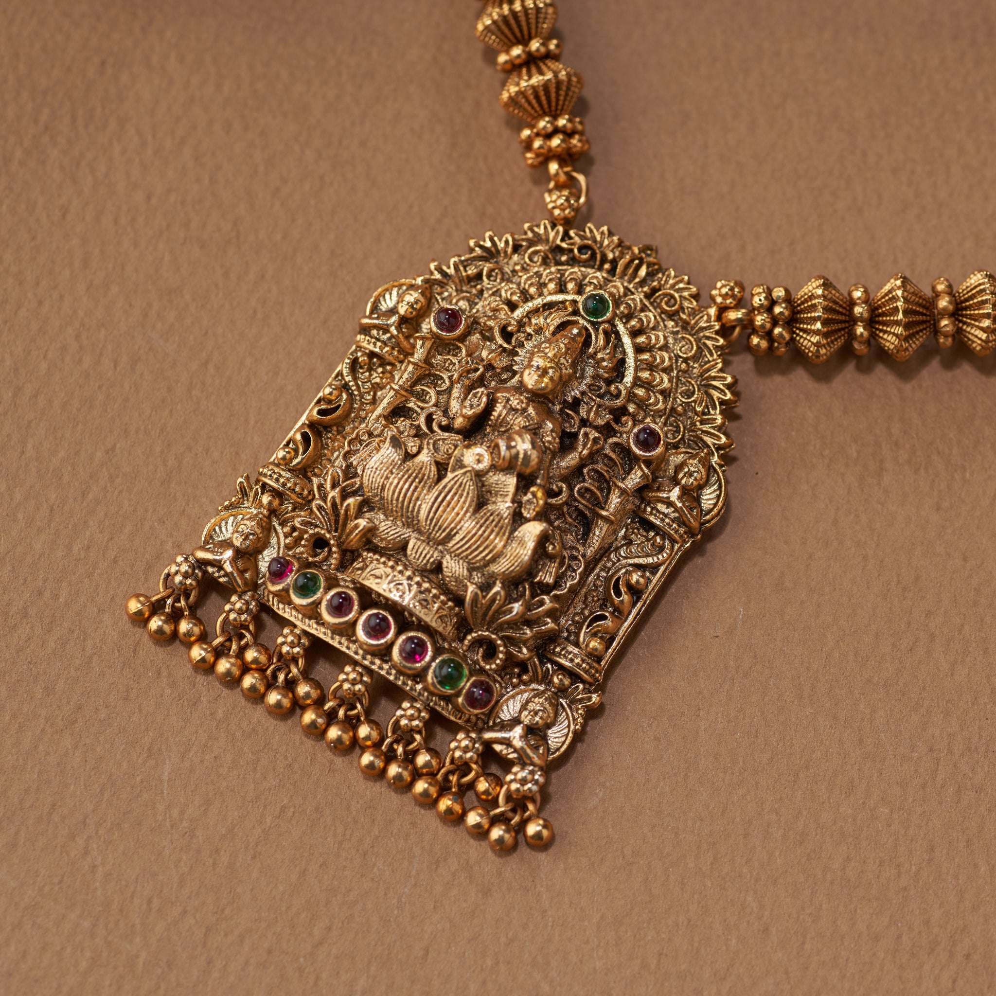 Gold necklace and earrings on a brown background. South Indian Temple jewelry with Lakshmi Motif. Temple Jewellery Lakshmi | Tilloma. Matt gold with intricate design on a brown background