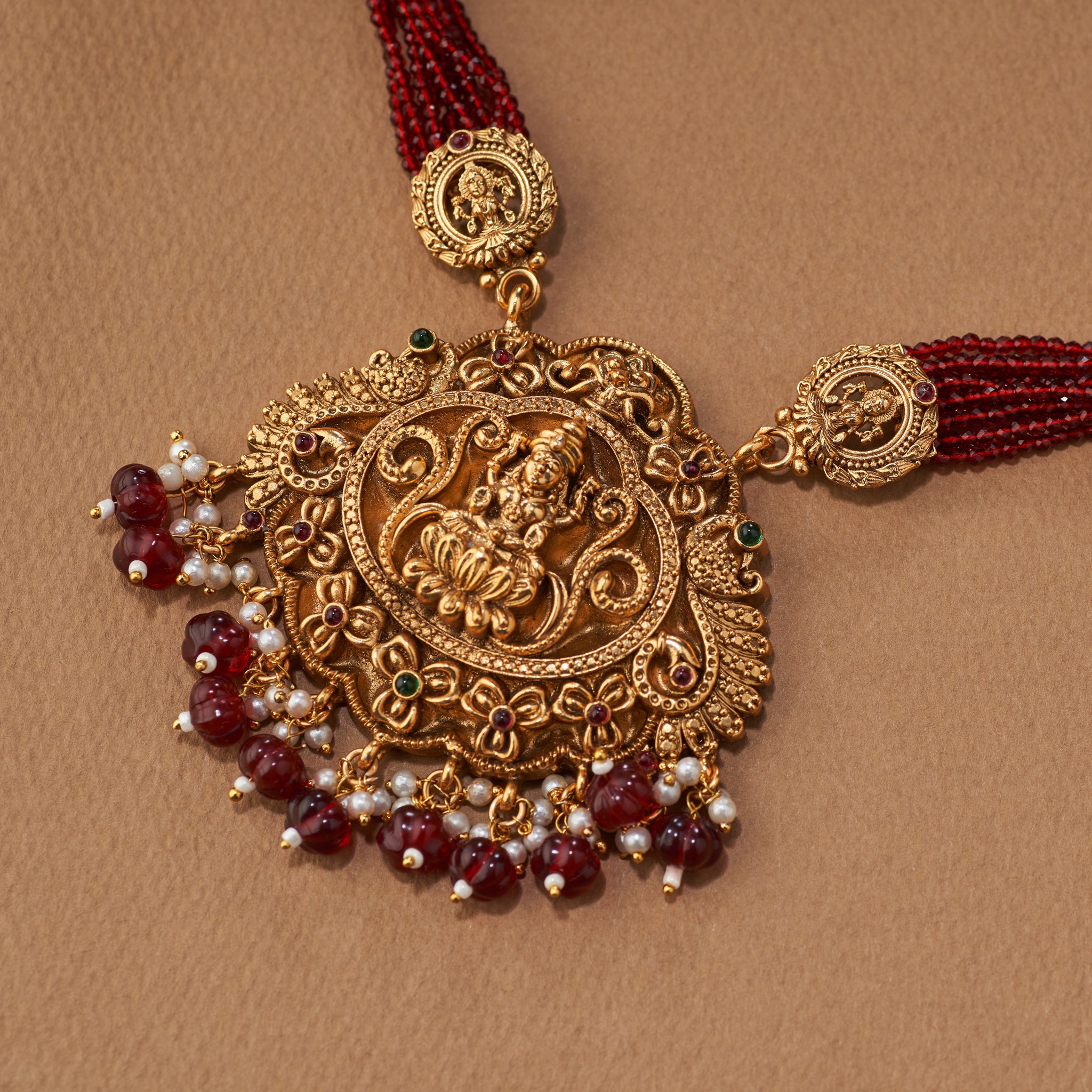 Gold necklace with red beads and tassels on a brown background. Maroon beaded gold Lakshmi pendant intricate design South Indian Temple jewellery necklace set with matching jumkhis or earrings. Matt gold plated.