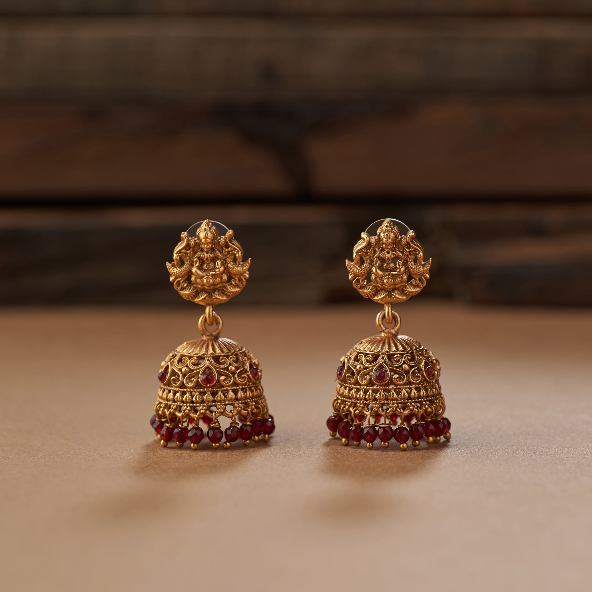 Pair of gold earrings with maroon gemstones on a brown background. Ornate gold necklace with marron beads on a brown background. Gold and maroon beaded necklace with matching earrings on a brown background. Maroon beaded necklace with gold Lakshmi pendants, South Indian Temple jewellery on a brown background. matt gold finish and paired with earrings.