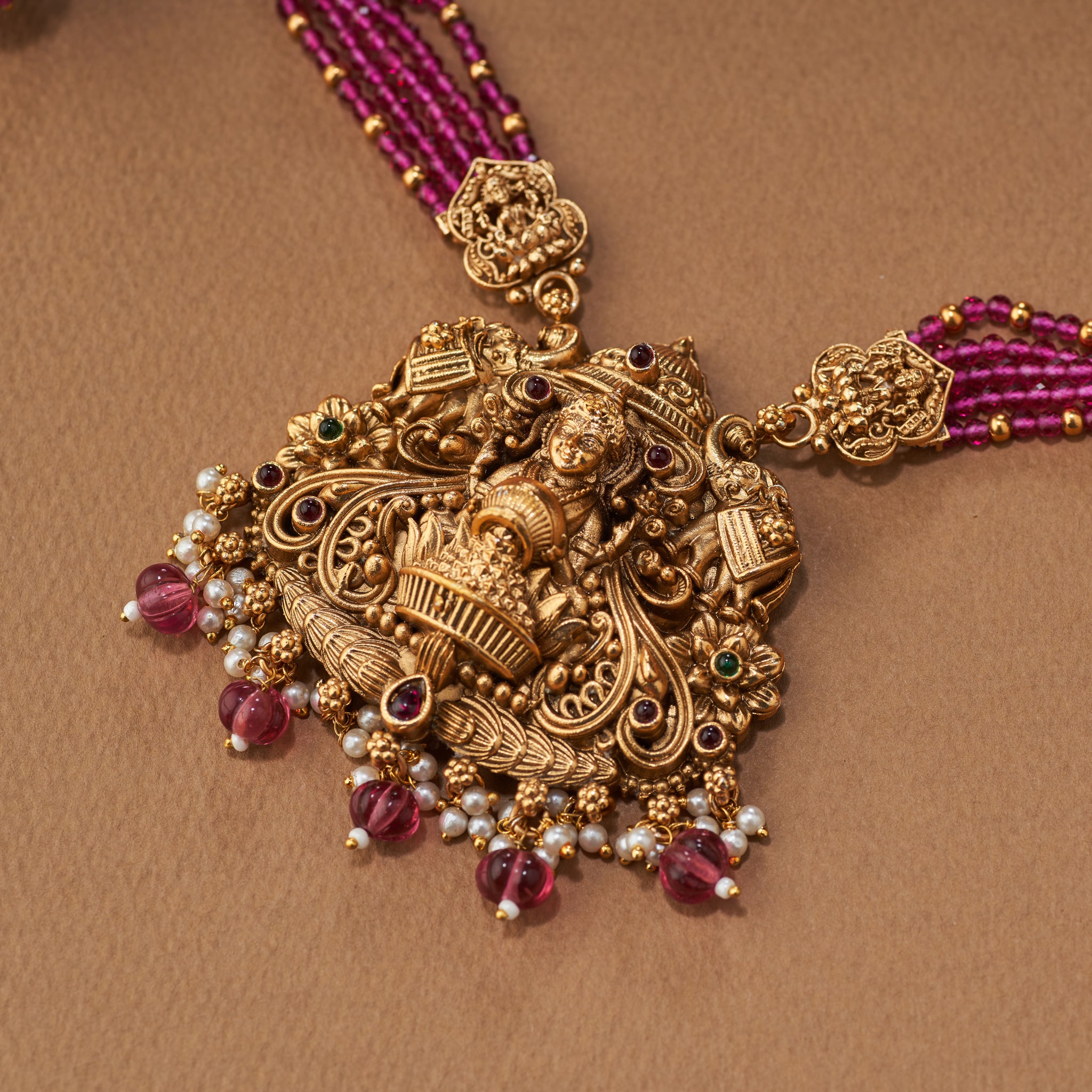 Decorative gold necklace with pink beads, Pink beaded necklace with gold Lakshmi pendants, South Indian Temple jewellery on a brown background. matt gold finish and paired with earrings.
