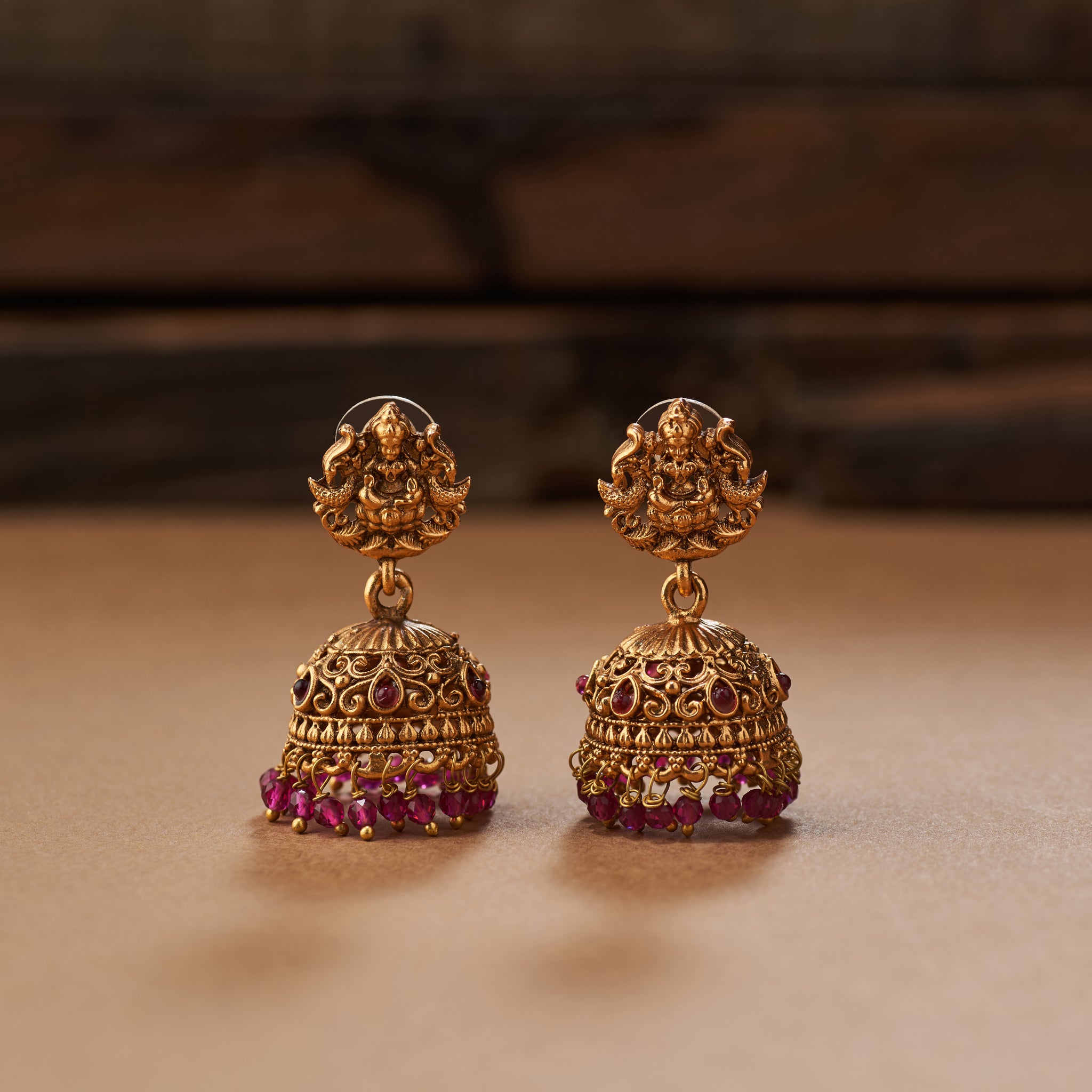 Pair of gold earrings with pink beads. Pink beaded necklace with gold Lakshmi pendants, South Indian Temple jewellery on a brown background. matt gold finish and paired with earrings.