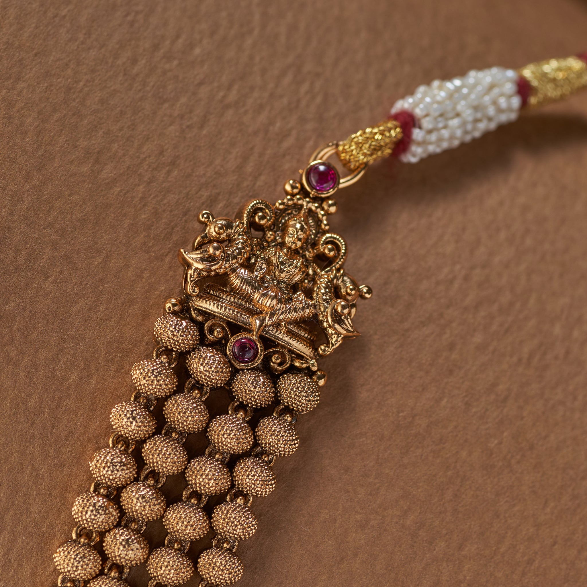 Close-up of an antique gold temple jewelry necklace pendant showing a detailed depiction of the goddess Lakshmi and peacocks with a ruby stone accent.