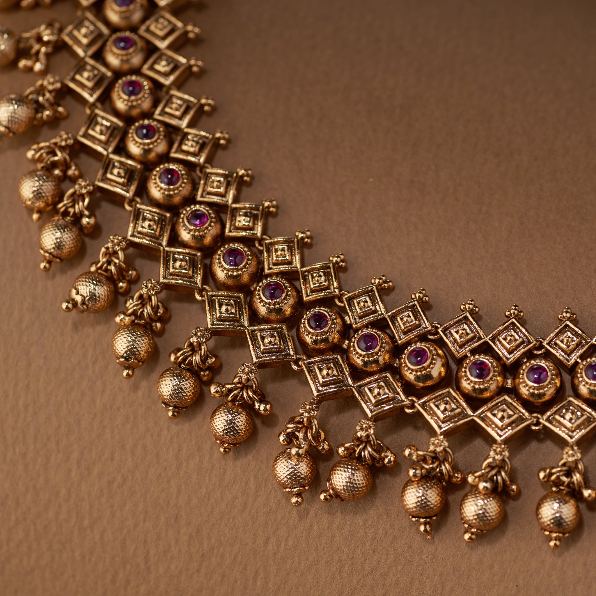 A close-up shot of an antique gold temple jewellery set, featuring a necklace and matching earrings with ruby stones and hanging gold beads, displayed on a textured surface.