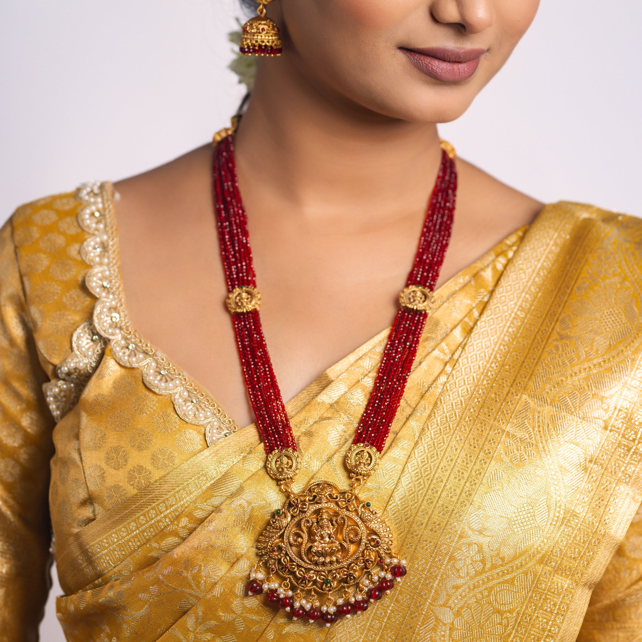Woman wearing a gold saree with a maroon beaded south Indian Temple necklace and earrings on a white background