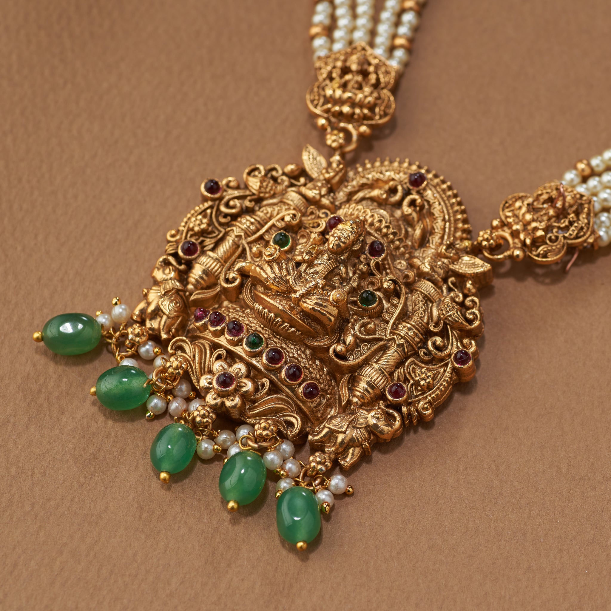Gold necklace with intricate design and green gemstones on a brown background, Ornate gold necklace with green gemstones on a brown background, South Indian temple jewellery with Lakshmi pendant, paired with matching earrings. Matt gold finish.