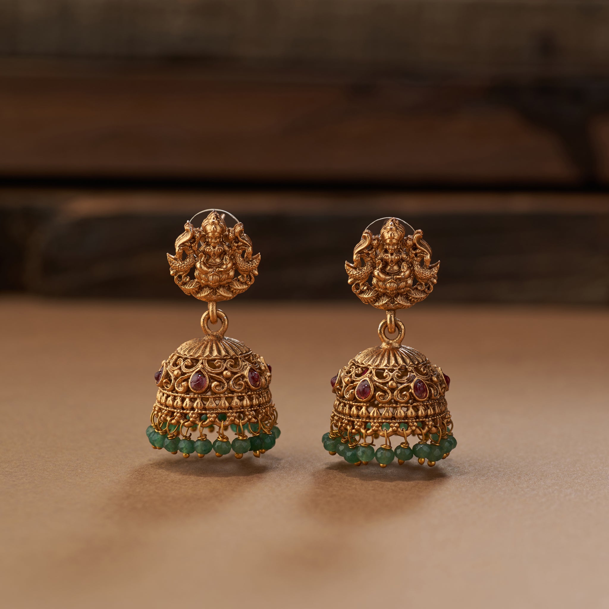 Gold earrings with green gemstones on a brown background. Gold south Indian template necklace with Lakshmi pendant and with off-white and green beads and matching earrings on a brown background. Matt gold plated.