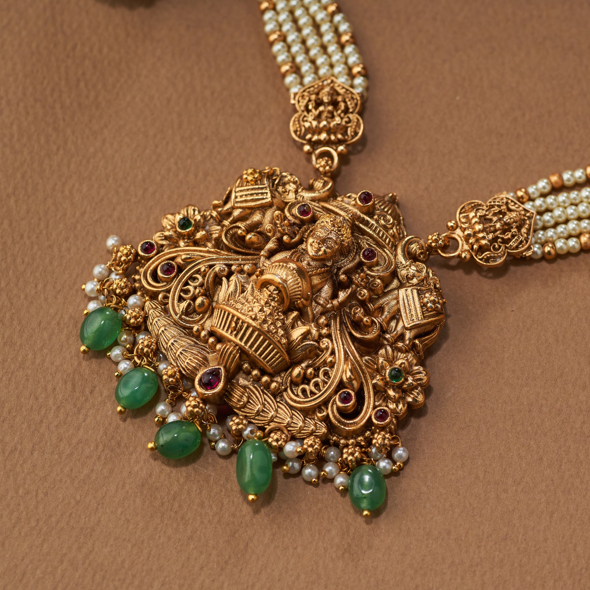 Ornate gold necklace with green gemstones on a brown background. Ornate gold necklace with green gemstones on a brown background, South Indian temple jewellery with Lakshmi pendant, paired with matching earrings. Matt gold finish.
