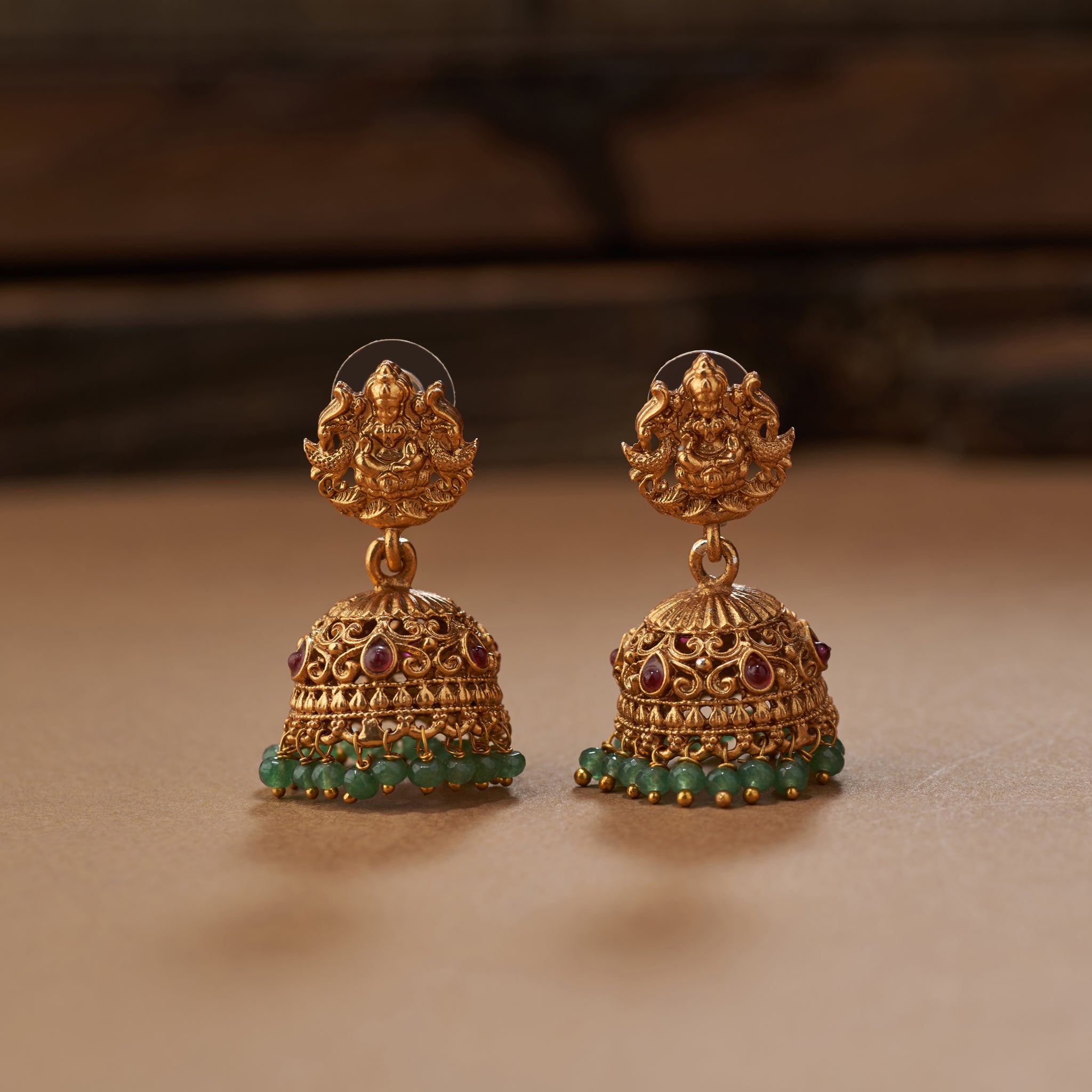 Pair of gold earrings with green beads on a brown background. Ornate gold necklace with green gemstones on a brown background, South Indian temple jewellery with Lakshmi pendant, paired with matching earrings. Matt gold finish.