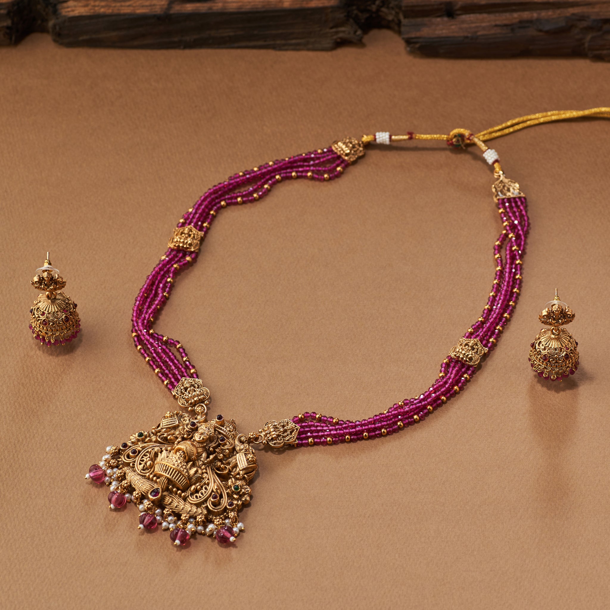 Pink beaded necklace with gold Lakshmi pendants, South Indian Temple jewellery on a brown background. matt gold finish and paired with earrings.