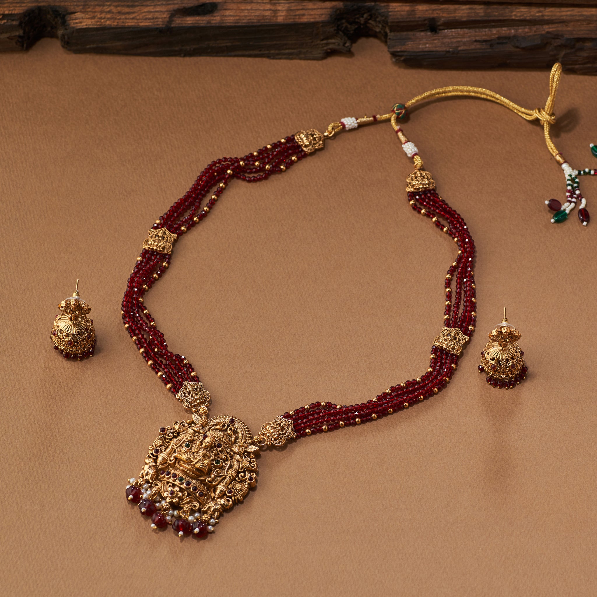 Gold and maroon beaded necklace with matching earrings on a brown background. Maroon beaded necklace with gold Lakshmi pendants, South Indian Temple jewellery on a brown background. matt gold finish and paired with earrings.