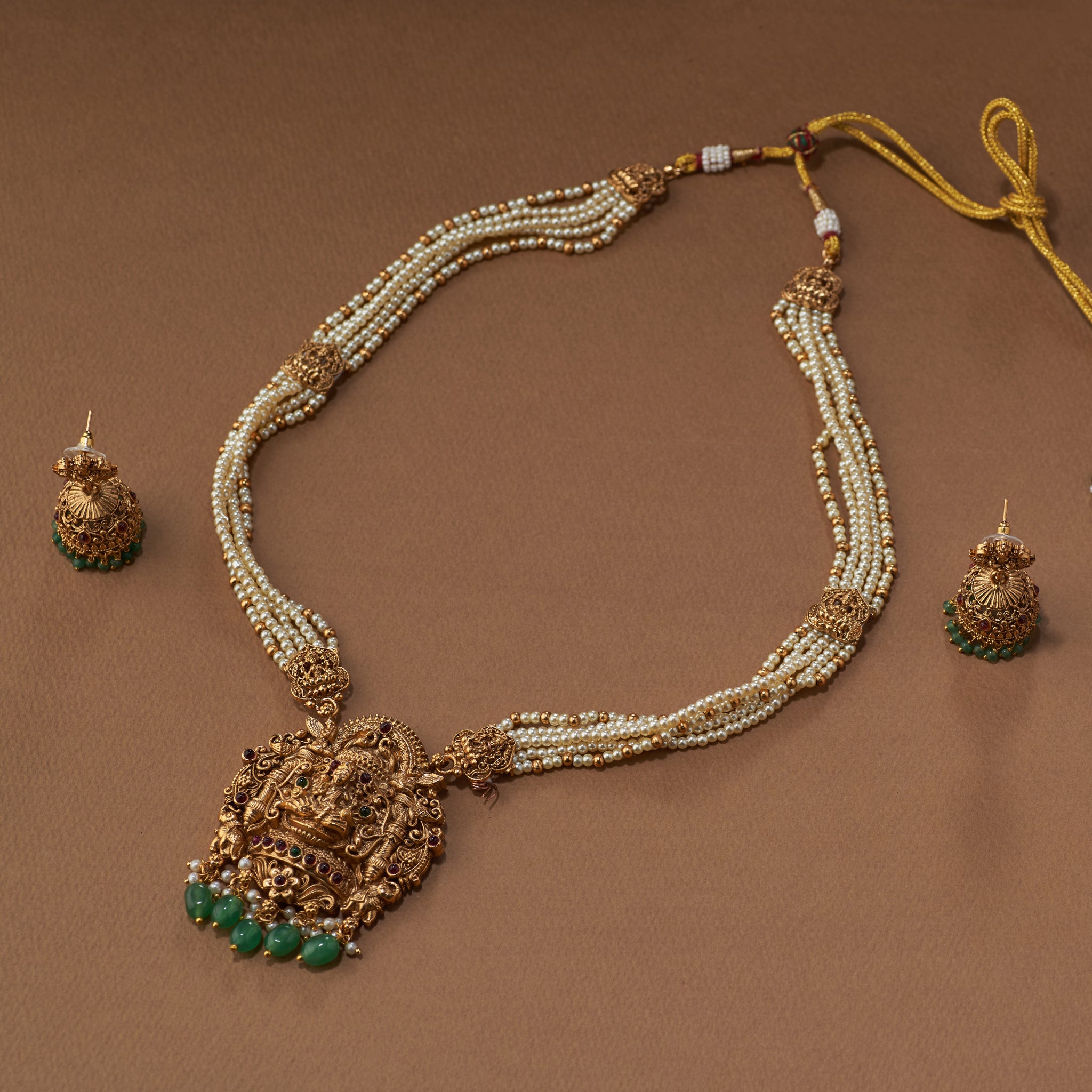 Gold south Indian template necklace with Lakshmi pendant and with off-white and green beads and matching earrings on a brown background. Matt gold plated.