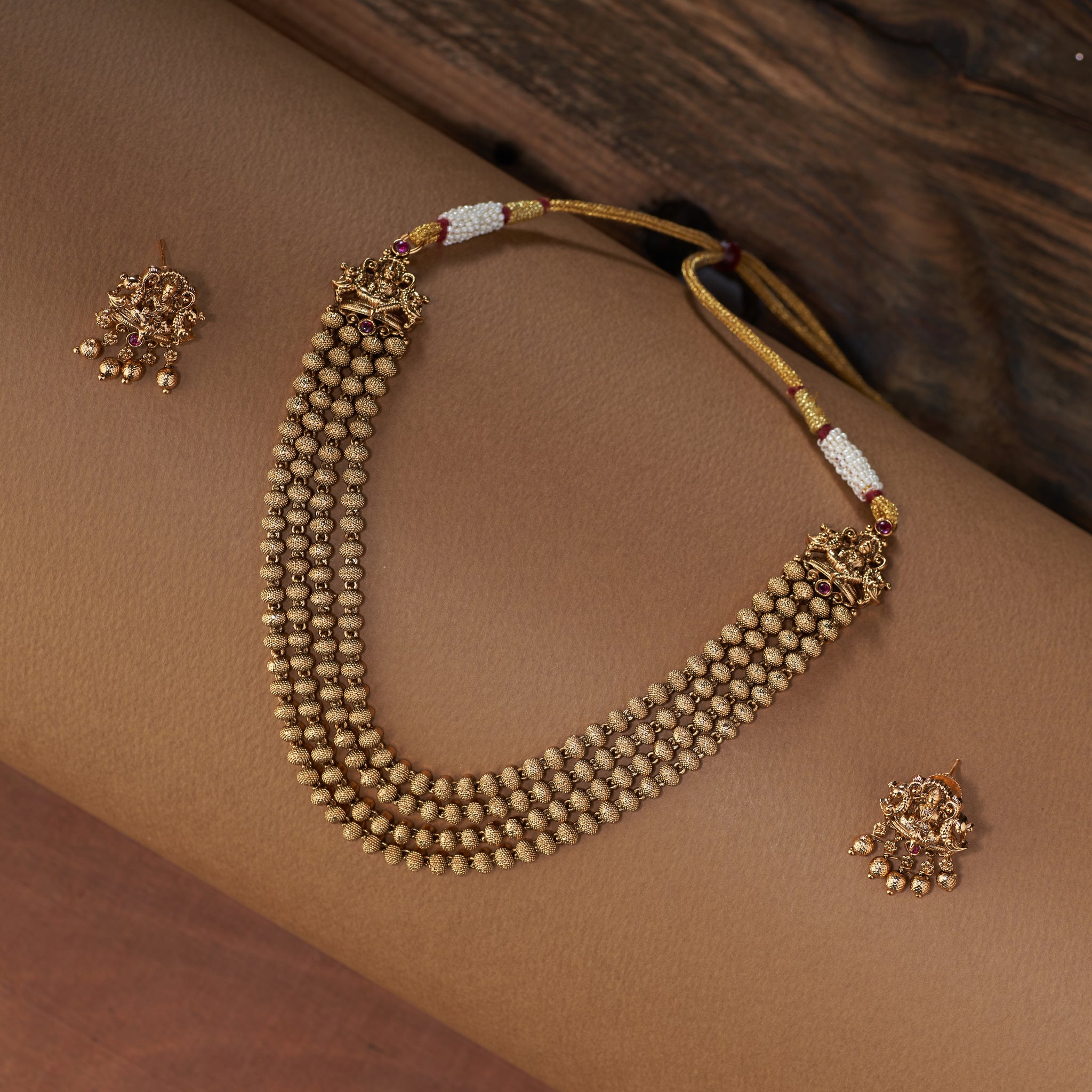 A full shot of the Tilloma antique gold temple jewelry set, featuring a four-layered beaded necklace with a Lakshmi pendant and matching stud earrings, displayed on a brown backdrop.