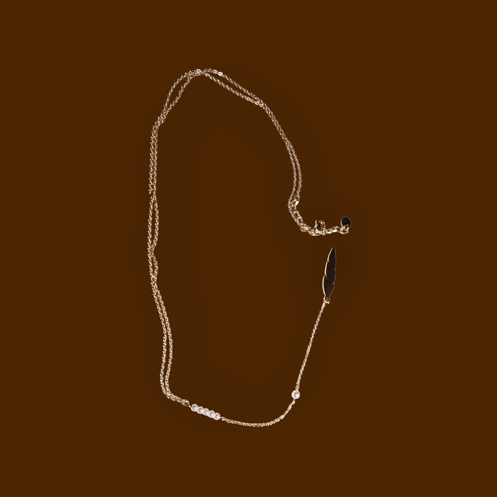 A vertical product shot of a long, delicate gold lariat necklace featuring a feather pendant and a row of small white pearls.